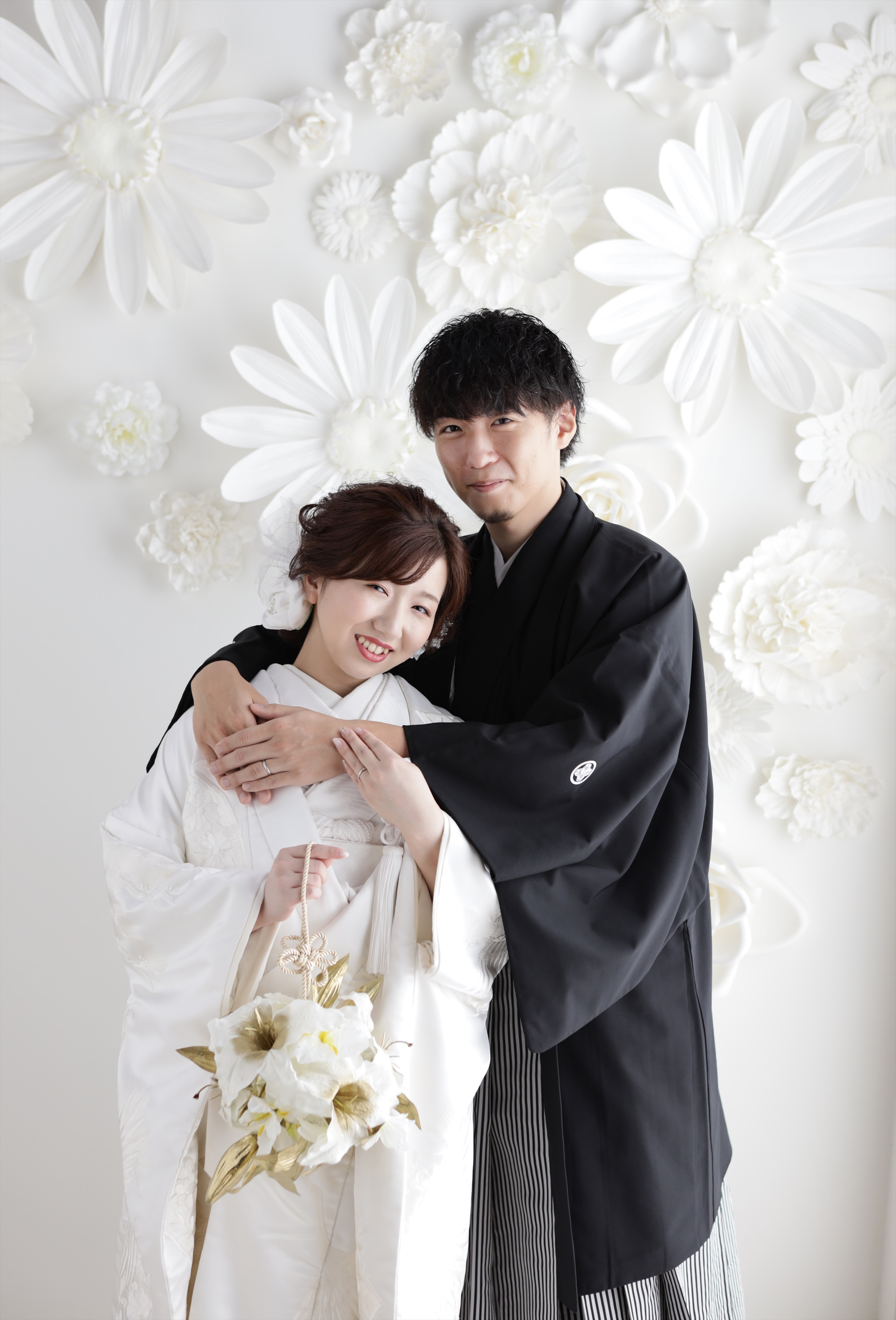 白い花の壁を背に寄り添う白無垢姿の新婦と黒紋付袴の新郎