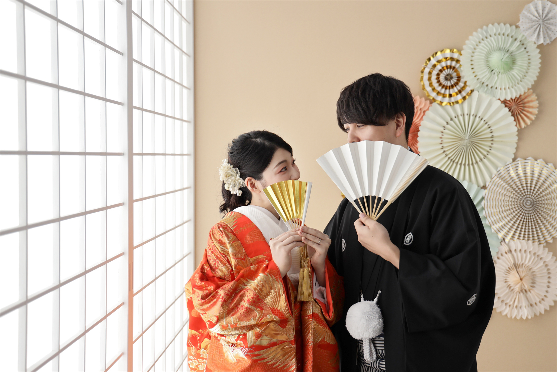 障子の光の中で末広を手に見つめ合う朱色の色打掛の新婦と黒紋付袴の新郎