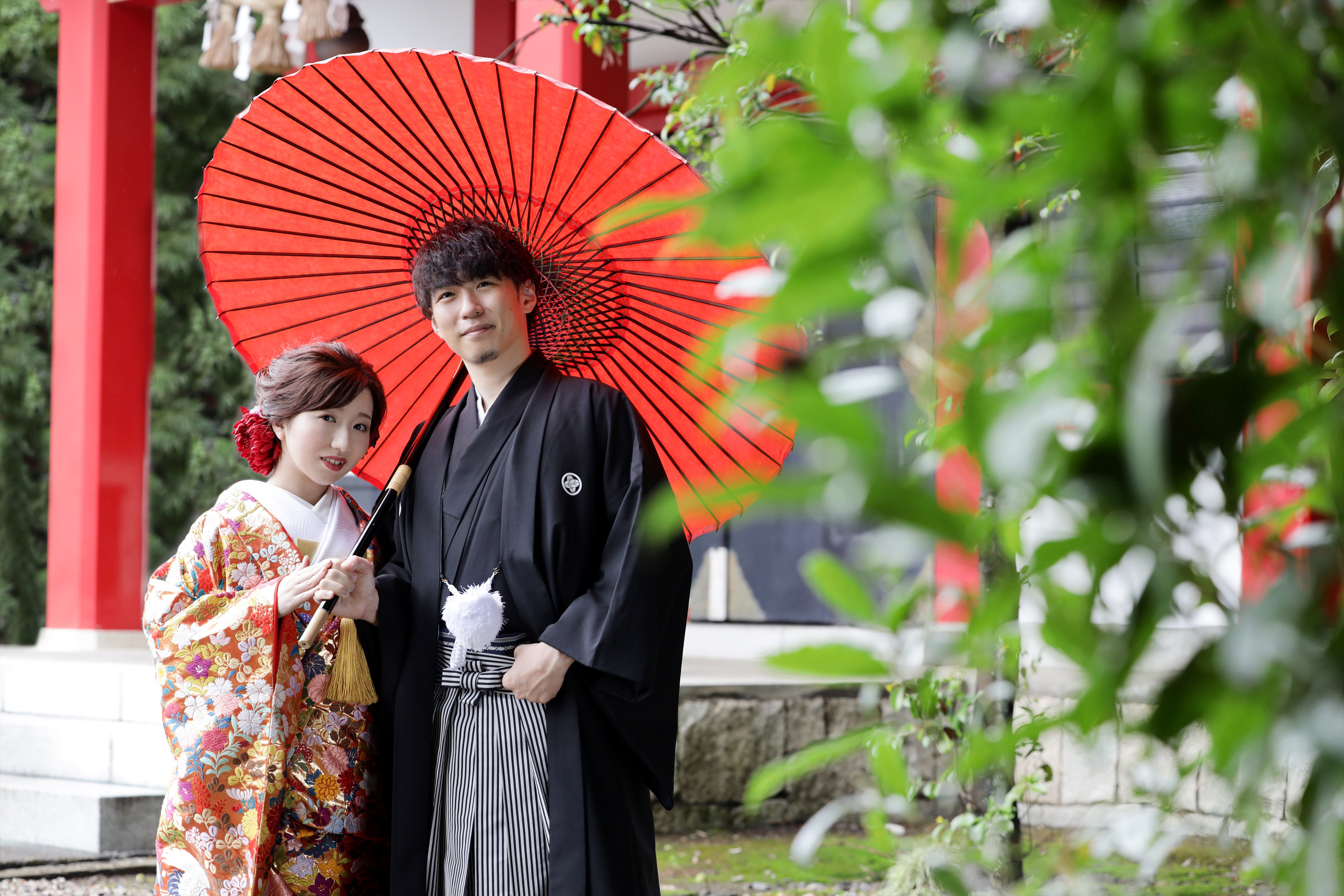 朱塗りの社殿前で赤い和傘をさす色打掛の新婦と黒紋付袴の新郎