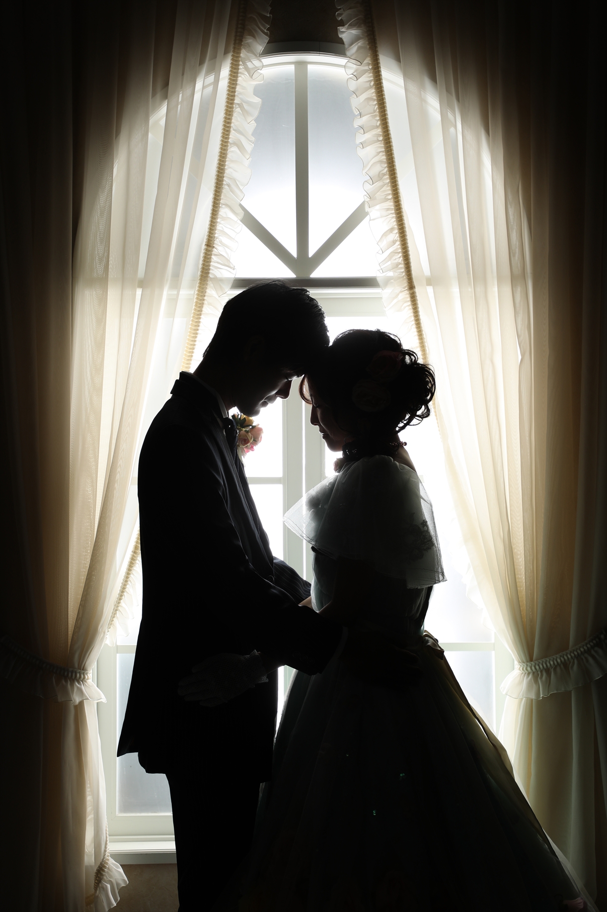 窓辺の逆光に浮かぶドレス姿の新婦とタキシードの新郎の寄り添うシルエット